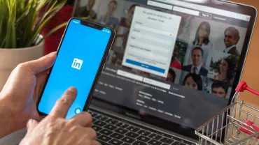 mão segurando celular logado no LinkedIn, notebook logado em LinkedIn ao fundo