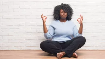 Mulher negra meditando, parede branca ao fundo.