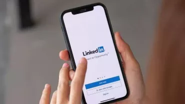 Mãos brancas segurando um celular que está com a página do LinkedIn aberta. 
