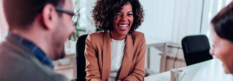 Três pessoas sorrindo em uma reunião de trabalho.