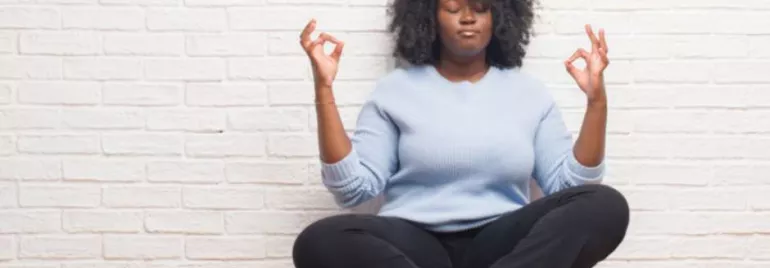 Mulher negra meditando, parede branca ao fundo.