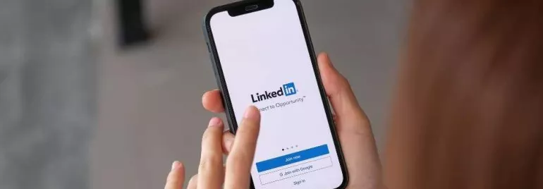 Mãos brancas segurando um celular que está com a página do LinkedIn aberta. 
