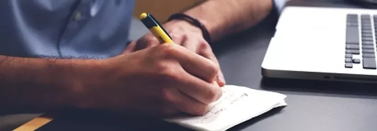 Pessoa escrevendo em um bloco de notas ao lado de um laptop.